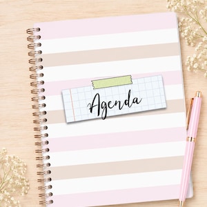 May include: A spiral-bound agenda notebook featuring horizontal stripes in alternating shades of light pink and beige. A rectangular piece of grid paper with the word Agenda written in black script is attached to the center with green tape.