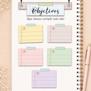 May include: A spiral-bound notebook page featuring a header with the text "Objetivos" and "Que deseo cumplir este a&ntilde;o" above five numbered, pastel-colored sticky note sections in yellow, beige, pink, green, and purple, each secured with tape.