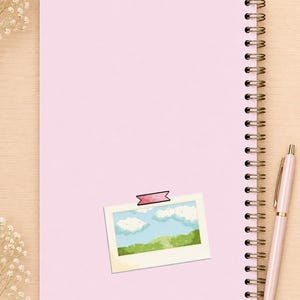 May include: A light pink spiral-bound notebook with a rectangular sticker featuring a pixel art landscape of a blue sky with white clouds and green hills, secured by a small pink tape graphic, accompanied by a matching light pink ballpoint pen.