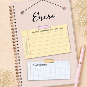 May include: A spiral-bound planner with a light pink cover featuring the word Enero in black script. It includes a yellow lined checklist titled Lo m&aacute;s importante para este mes and a white grid note section labeled Para Inspirarme, accompanied by a pink pen.