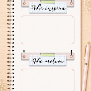 May include: A spiral-bound notebook page featuring two rectangular sections with pink borders. Above each section is a grid-paper label attached with pins and tape, reading Me inspira and Me motiva in black script, accompanied by a light pink pen.