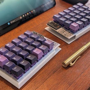 Könnte beinhalten: Zwei benutzerdefinierte mechanische Tastaturen mit lila und schwarzen Tastenkappen, ein Messingstift und ein Tablet auf einer Holzoberfläche. Die Tastaturen haben ein kompaktes Layout, und die Tastenkappen sind auf einer weißen Basis angeordnet.