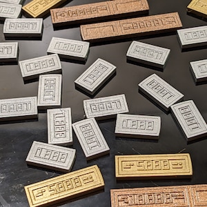 Op de afbeelding: Een assortiment rechthoekige metalen objecten in goud, zilver en brons. Elk stuk heeft het nummer "5888" of "1888" in het oppervlak gegraveerd. De objecten liggen verspreid op een donkere, reflecterende ondergrond.