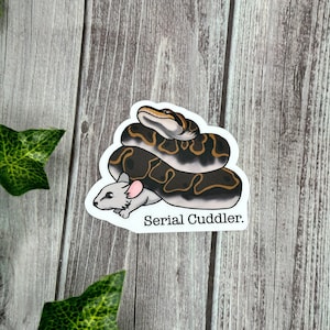 May include: A sticker featuring a cartoon illustration of a snake coiled around a mouse. The snake is brown and black with yellow accents. The mouse is gray with pink ears. The text "Serial Cuddler" is below the mouse.
