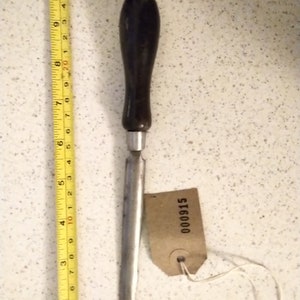 Puede incluir: Una lezna vintage con un mango de madera oscura y un eje de metal plateado. La lezna mide aproximadamente 15 cm de largo. Una pequeña etiqueta con el número 000915 está adjunta. Una cinta métrica amarilla está al lado de la lezna.