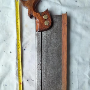May include: A vintage hand saw with a wooden handle and blade. The handle is a dark brown color and is attached to the blade with brass rivets. The blade is a rectangular shape and is approximately 15 inches long. A yellow measuring tape is next to the saw.