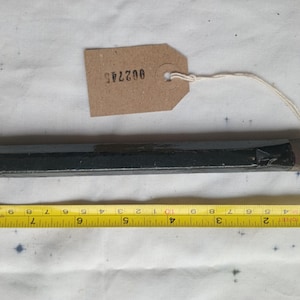 Pode incluir: Um cinzel preto e castanho com uma lâmina de metal, com aproximadamente 20 cm de comprimento, com uma fita métrica amarela por baixo. Uma etiqueta castanha com o número "002745" está presa ao cinzel.