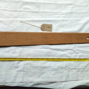 May include: A wooden framing square with a long arm and a shorter arm, on a white surface. A yellow measuring tape is placed below the square. The square has a small tag with the number "PA2032" attached. The tape shows measurements in cm.