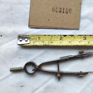May include: A metal drafting compass with a knurled handle and adjustment screws, positioned below a yellow measuring tape showing a length of approximately 7.6 cm and a brown rectangular paper tag with the printed number 003180.
