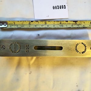May include: A brass spirit level measuring approximately 18 cm in length, positioned below a yellow measuring tape. The tool features the text "ELVICTA", "525", and "WARRANTED CORRECT" engraved on its surface, along with a central glass vial.