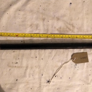 May include: A black, rectangular object, possibly a tool, next to a yellow measuring tape. The tape shows measurements in inches. A small, brown tag with text is attached to the object with a string. The object appears to be about 12 inches long.