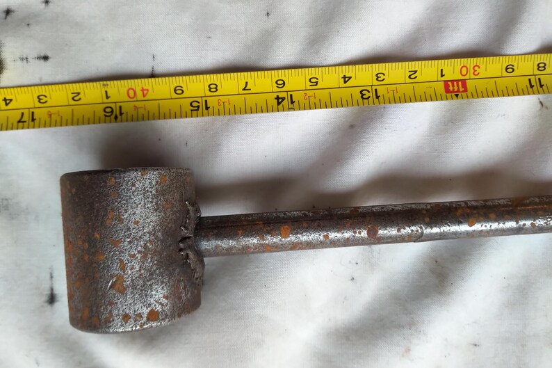 Peut inclure: Un outil en m&eacute;tal rouill&eacute; avec une t&ecirc;te cylindrique et une longue poign&eacute;e. Un ruban &agrave; mesurer jaune est plac&eacute; &agrave; c&ocirc;t&eacute; de l'outil, indiquant des mesures. L'outil mesure environ 35,5 cm de long.