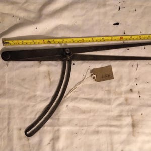 May include: A vintage, dark metal caliper tool with curved handles and pointed tips, measuring approximately 30 cm in length. A yellow measuring tape is placed alongside the tool. A small tag is attached.