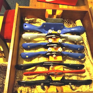 May include: A wooden box filled with various woodworking tools, including several drawknives in blue, black, and red. The tools are arranged on a bed of yellow packing material. A red Nike shoe box is visible in the background.