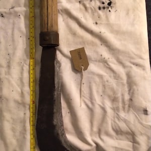 May include: An antique curved blade tool with a wooden handle and metal collar. A yellow measuring tape is placed next to the tool. A small tag with the number "002051" is attached to the tool.