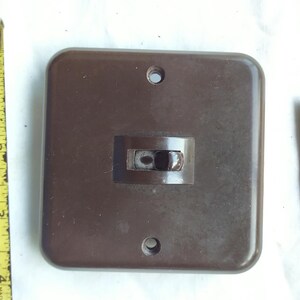 Puede incluir: Un interruptor de luz cuadrado de color marrón con una palanca central y dos orificios de montaje. Una cinta métrica amarilla muestra que el interruptor mide aproximadamente 9 cm de ancho. Una pequeña etiqueta beige tiene el texto 4003176.