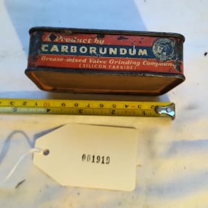 May include: A rectangular metal tin of Carborundum grease-mixed valve grinding compound. The tin is red and black with the product name and description printed on it. A yellow tape measure is next to the tin, and a white tag with the number 001919 is attached.