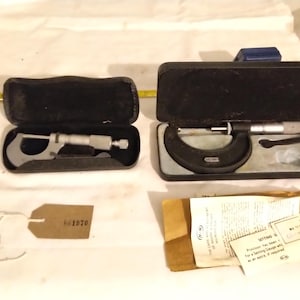 May include: Two precision micrometers in open black cases. One is a larger, black micrometer, the other is smaller and silver. A brown tag with the number 661970 is attached to a white string. Documents are also present.
