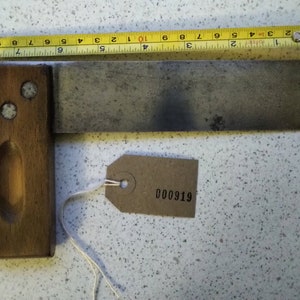 Op de afbeelding: Een houten en metalen winkelhaak met een bruine houten handgreep en een metalen blad. De handgreep heeft drie metalen klinknagels. Een bruin papieren label met het nummer 000919 is bevestigd aan de winkelhaak.