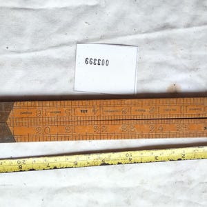 May include: A vintage wooden folding ruler with brass hinges and black numerical markings, positioned above a yellow metal measuring tape that shows a length of approximately 10 inches. A small white tag displays the black printed text 003399.