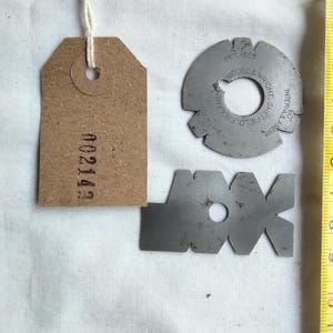 May include: A collection of vintage metal tools and a brown tag with the number "002143" printed on it. A yellow tape measure shows measurements in inches. The tools include a circular piece with text and a shape with the letters "JAX".