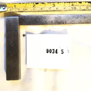 May include: A grey metal engineer's square positioned next to a yellow measuring tape that shows the tool is approximately 12.7 cm long. A white rectangular tag with the black printed text 0034 5 is placed near the centre of the metal square.