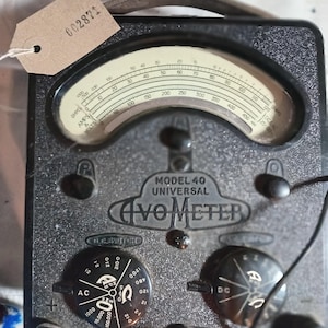 May include: Vintage Model 40 AVO Meter, a universal electrical measuring instrument. The black meter features a curved analog display with numerical markings and a needle. It has multiple dials, switches, and input terminals, including a red output cable. A tag with the number 002871 is attached.