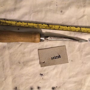 May include: A metal tool with a wooden handle positioned next to a yellow measuring tape showing a length of approximately 10 inches. A small rectangular tag with the printed number 003286 and a small blue plastic piece are also visible.