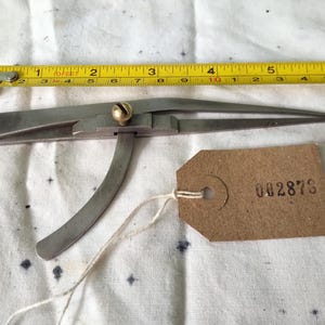 May include: A vintage metal caliper tool with a brass screw and a brown tag with the number 002873. A yellow tape measure is placed next to the caliper. The tools are on a white surface.