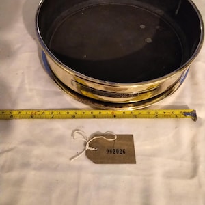 Może przedstawiać: Mosiężne sito z ciemnym wnętrzem, niebieskim paskiem i złotą obwódką. Żółta taśma miernicza znajduje się obok sita. Do sita przymocowana jest brązowa etykieta z numerem "002026".