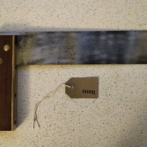Op de afbeelding: Een houten en metalen winkelhaak met een messing rand en drie messing schroeven. De metalen blad is licht aangetast. Een bruin papieren label met het nummer 000931 is bevestigd aan de winkelhaak.