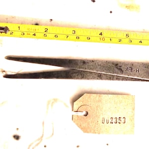 May include: A metal caliper tool with the text "A-E-H" and a tag with the number "002353". A yellow measuring tape shows measurements in inches. The caliper is approximately 15 cm long.