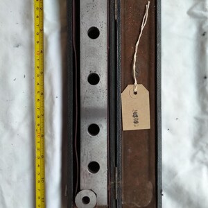 May include: An open, vintage, black leather case with a metal tool inside. The tool is a rectangular bar with five circular holes and two circular ends. A yellow measuring tape is next to the case. The tool is approximately 25 cm long.