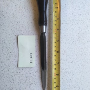 May include: A wood carving tool with a black handle and a metal blade, measured against a yellow tape measure. The tool is approximately 20 cm long.