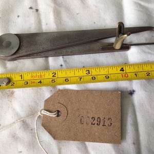 May include: An antique metal tool with a brass accent, a yellow measuring tape showing centimetres, and a brown tag with the number "002913". The measuring tape shows up to 15 cm.