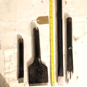 May include: A collection of black chisels with silver edges and brown handles, ranging in size. A yellow tape measure is next to the chisels. A small tag with text is attached to one chisel.