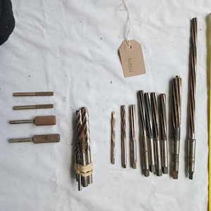 May include: A selection of metal tools, including reamers and drill bits, laid out on a white surface. A yellow measuring tape is placed beside the tools. A brown tag with the number "012153" is attached to the tools.