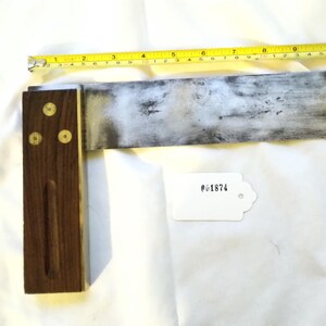 Könnte beinhalten: Ein alter Zimmermannswinkel mit einem dunklen Holzgriff und einer Metallklinge. Die Klinge weist Gebrauchsspuren auf. Ein gelbes Maßband liegt neben dem Winkel. Ein weißes Etikett mit der Nummer #01874 ist ebenfalls vorhanden.