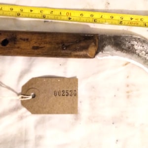 Op de afbeelding: Een vintage mes met gebogen lemmet en houten handvat, ongeveer 15 cm lang, en een metalen lemmet. Een gele rolmaat ligt naast het mes. Een bruin label met het nummer 002536 is ook aanwezig.