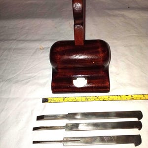 May include: A dark brown wooden woodworking plane with a vertical handle and a central opening, accompanied by three flat metal blades. A yellow measuring tape is positioned below the plane, showing a length of approximately 23 cm.