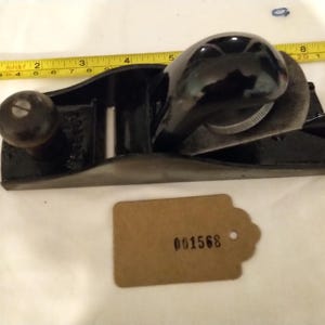 以下が含まれることがあります： 木製のハンドルと金属製の刃が付いた黒い手押し鉋。鉋には「Record」と表示されており、横にメジャーが置かれている。