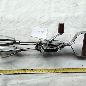 Könnte beinhalten: Ein manueller Handmixer mit zwei Metallquirlen, einem verchromten Getriebemechanismus und zwei braunen Holzgriffen. Ein gelbes Maßband liegt unter dem Mixer und zeigt eine Gesamtlänge von etwa 34 cm. Ein kleines weißes Etikett mit der Aufschrift 003337 ist angebracht.