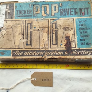 May include: Vintage "Tucker Pop Rivet Kit" box with instructions and illustrations. The box is aged with wear and tear, featuring blue and tan colors. A yellow measuring tape and a tag with the number "002680" are also present.