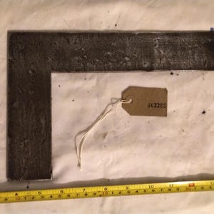 May include: A dark, L-shaped metal square tool with a textured surface. A small, brown tag with the number "J62285" is attached. A yellow measuring tape with black markings is at the bottom.
