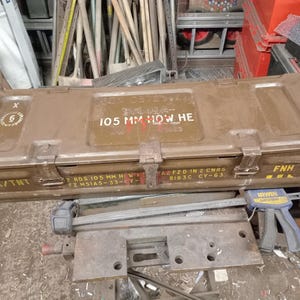Puede incluir: Una caja de municiones rectangular de metal verde oliva con el texto "105 MM HOW HE" y otras marcas. La caja tiene una tapa con bisagras y un pestillo y está etiquetada como "RDX/TNT".