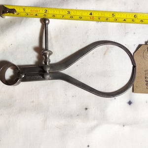 May include: A vintage metal caliper with a screw adjustment, next to a yellow measuring tape showing inches. A small brown tag with the number 006290 is attached to the caliper. The tools are on a white surface.