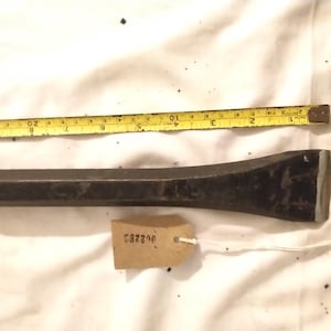 Puede incluir: Un cincel de metal oscuro con una hoja rectangular y un mango cilíndrico, junto a una cinta métrica amarilla. El cincel mide aproximadamente 20 cm de largo. Una pequeña etiqueta con el texto "C82200" está adjunta.