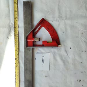 Może przedstawiać: Czerwona metalowa głowica kątomierza przymocowana do długiej stalowej linijki, umieszczona obok żółtej miarki Stanley z podziałką w calach i centymetrach. Dołączona jest mała biała kwadratowa etykieta z nadrukowanym numerem 003373.
