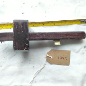 Puede incluir: Un calibrador de marcado de madera vintage con un acabado marrón oscuro, con una cabeza deslizante y un detalle de latón. Una cinta métrica amarilla está al lado del calibrador. Una pequeña etiqueta marrón con el número 002772 está adjunta.