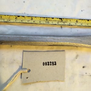 Puede incluir: Un cincel de metal plateado con eje hexagonal, colocado debajo de una cinta métrica amarilla que muestra una longitud de aproximadamente 20 cm. Una etiqueta de papel rectangular con el número impreso 003283 está unida a la herramienta.